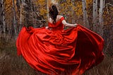 A woman in a red ball gown and gold crown, dancing in front of birch trees in the fall.