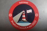 Close up on a sticker on a laptop lid. The sticker reads ‘Intranet Product Launch. Jul 2023’. The design on the sticker is a striped red and white lighthouse on a rock shining a light onto the sea. There is a flag with the Royal Borough of Greenwich logo on it next to the lighthouse, and two tudor rose emblems (also part of the borough’s logo) on either side of the sticker.