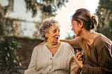 A woman talking with her elderly mother while smiling and putting an arm on the mother’s shoulder.