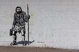 stencil work by thepinkbear.rebel street artist of a neanderthal man holding a briefcase in one hand and a spear in the other. Wearing a shirt, tie and suit jacket over an animal skin. Ready for a solid days work.