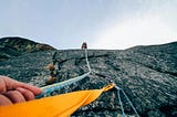 A hand holds onto a blue rope, the photographer attached to an orange harness, the camera looking up the face of a cliff, with another person far ahead, reaching the top
