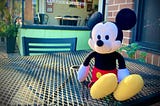 Image shows a stuffed Mickey Mouse doll sitting propped up on a patio table outside a restaurant window that says “Raw food and Desserts”