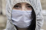 A young woman wears a face mask over her mouth & nose, as well as a protective hood over her head. She looks into the camera.