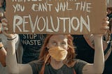 Photo of a young woman in a crowd of protesters marching together. She is wearing a COVID mask and holding up a cardboard sign with the hand painted message, “YOU CAN JAIL THE REVOLUTIONARY, BUT YOU CAN’T JAIL THE REVOLUTION”