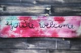 Photograph of a tie dyed piece of fabric with the words “You’re welcome” stitched on it. It was ironed onto a cloth mask.