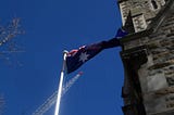 Australian Flag flying against blue sky with an old sandstone building and building crane in background.