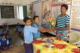 Image is from the event Samman Samaroh. The image shows a student standing on the left handing over a bouquet to a staff