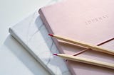Two pencils with pink tips sit on top of a pink journal with the words JOURNAL written in the middle.