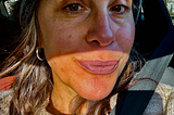 Photo of woman in hat with gray hair. Half her face is in a shadow, mouth in the light, half smiling