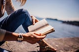 Photo of a woman reading a book looking out over a lake