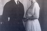 Felix and Irina standing together. He’s in a dark suit with short, slicked-back hair. She’s in a white day dress with pearls.