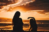 A woman and a young girl sitting on the beach, facing the sunset with their backs to us
