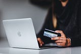 A person holds a credit card near a laptop, appearing to type in information to purchase something online.