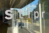 Window with “s t l p r” logo in white lowercase letters (for the St. Louis NPR-affiliate station) reflects a street scene outside with a blue sky with a few white clouds. Inside, one can glimpse some framed art from a gallery in the lobby. (Photo by author.)