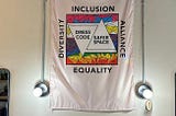Dress Code Project flag — White Flag hung down from the ceiling with a square box with Inclusion on top, Alliance to the right, Equality at the bottom and Diversity on the left. Inside the box is rainbow colors plus pinks and whites and two triangles one upside down with the words Dress Code Safer Space.