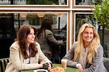Two women are sitting at a bistro table drinking coffee.