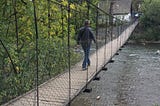 A man wearing a leather jacket and denim jeans walks across the boards of a suspension bridge over a river. The bridge is narrow with chain-fence sides, and trees are at the edge.