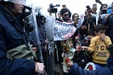 Refugee children offer flowers to riot police after clashes outside a refugee camp in the village of Diavata, west of Thessaloniki, northern Greece, Saturday, April 6, 2019. Clashes between hundreds of protesting migrants and police continue for a third day outside an overcrowded migrant camp, with migrants throwing rocks at police and the latter responding with tear gas and stun grenades. (AP Photo/Giannis Papanikos)