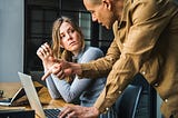 A woman with arms resting on a desk listens to a man standing next to her, pointing at a laptop screen.