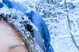 Half of Tabitha’s face, blue eye looking skyward wearing a blue hood as snowflakes fall around — she looks happy