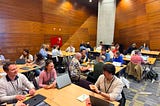 A room full of people engaged in group discussions around tables, using laptops and taking notes.