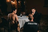 A man and a woman at a restaurant table, speaking to a man wearing a director’s shirt, back to the camera.