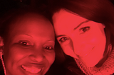 Photo of Sonja and Kristine smiling up to a camera. The image is filtered in red.