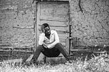 A black man in front of an old shack sitting at door with one hand on his head