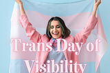 Woman holding Trans Pride Flag with great green and brown hair, a pink jacket and light blue shirt. The words Trans Day Of Visibility overlaid on top