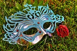 Red rose beside a blue sparkly Mardi Gras mask lying in the grass.