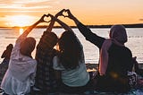 Four women sitting in front of a body of water at sunset, holding their hands together to create the shape of two hearts.