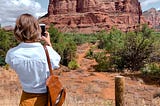 En route to Sedona, Arizona, one female tourist takes a moment to capture the natural beauty with her camera phone. She is a temporary visitor on the left in the photo. Next to her, on the right, a waist height pole supporting a barbed wire fence prevents temporary visitors from going any further. It is a permanent fixture. Photo is edited in color with a warm tone and cropped 16 x 9.