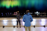 Two people wearing VR headsets stand on a balcony with an Aurora floating in the sky above the London skyline.