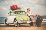 A woman leans on a Volkswagen Beetle parked on the beach. She has several pieces of luggage with her.