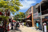 A street in Playa del Carmen, Mexico. The street is lined with restaurants and shops, and brightly colored flags hang overhead. Tourists and locals stroll each way, and the shining sun gives everything a vivid look.