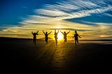 A group of people running on sand towards a sunset, leaping into the air