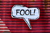 A white comic speech balloon containing the word “FOOL!” It appears to be on a red wall.