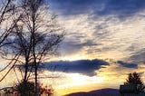 Sun tires to break through beneath dark gray and white clouds with bare winter tree in foreground.