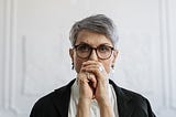 A woman in her late 50s/early 60s stairs straight at the camera looking stern with her hands folded in front of her mouth.