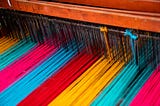 In the photo is a loom. It has thousands of threads in blocks of colour from yellow, aqua, pink, blue, orange, yellow, aqua, pink. The loom is wooden.