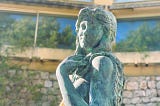 Top half of a stone statue of a naked young woman. Her arm covers her chest area.