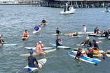 Surfers, swimmers and paddle boarders gather around a woman reading a poem elegy on her surfboard in a bay with a pier behind them.