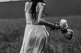 Black and white photo of a woman walking through tall grass in the rain, holding a bouquet. Her back is turned and her hair is wet, creating a moody and distant atmosphere.