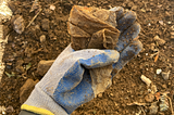 A gloved hand holds a piece of plastic waste, heavily soiled and degraded, underneath the thumb. The background is of dirt and soil in a garden.