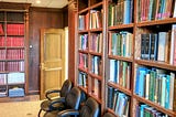 Library room with tall wooden bookshelves filled with neurosurgery and neuroscience books. Left shelf holds uniform red journals. Four black office chairs line the shelves. An open wooden door leads to a hallway. Room is warmly lit and quiet.