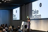 Black woman standing at a podium on on a conference stage. Two screen behind her read ‘Take action’