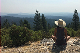 A brown-skinned woman sits on a rocky dirt area overlooking a vast expanse of evergreens. She wears a wide-brimmed hat and dark clothing.