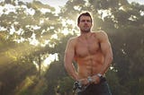Screenshot taken from The Diet Coke Ad on You Tube. Showing a man set against trees with piercing sunshine rays, he has his top off and is ringing it.