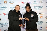 This images shows Stephen Arnott and David Okwesia from Beats Bus collecting the Hall of Fame award. They’re both holding the trophy between them infront of a background with sponsor logos