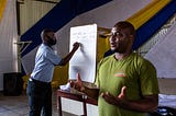 A man in a green shirt talks while a man in a blue shirt writes on a white board.